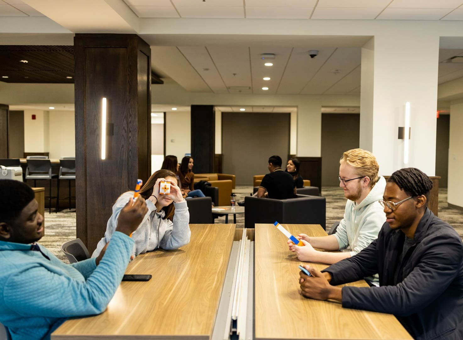 A group of students sits at a long table in a study room working on models. One student has made a viewfinder and looks straight at the camera.